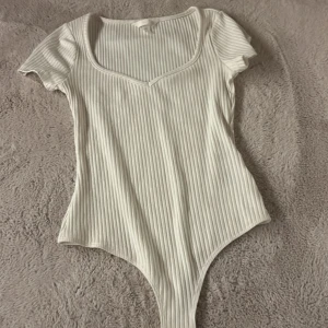 Vit ribbad bodysuit från H&M - Säljer en vit ribbad bodysuit från H&M med korta ärmar och djup rundad ringning. Bodyn är i stretchigt material och har en tight passform. Jag säljer denna då jag inte kommer använda den och är aldrig använd priset kan diskuteras💗