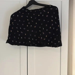 Svart blommig kortkjol från H&M Divided - Supersöt svart kortkjol från H&M Divided med små blommiga mönster i olika färger. Kjolen har omlottknytning i sidan och är tillverkad i ett lätt och luftigt viskostyg. Perfekt för dig som gillar en feminin och trendig stil.