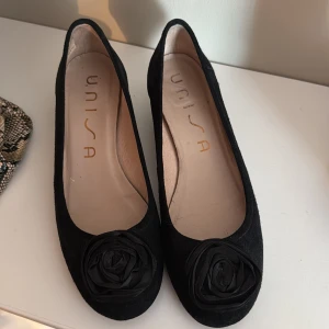 Svarta ballerinaskor med blomdetalj från Unisa - Säljer ett par svarta ballerinaskor från Unisa med rund tå och en dekorativ blomma framtill. Skorna har en klassisk och stilren design, perfekta för dig som gillar enkla men snygga detaljer.
