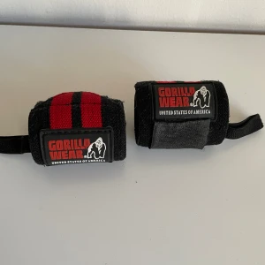 Svarta och röda träningshand wraps från Gorilla Wear - Säljer ett par svarta och röda hand wraps från Gorilla Wear, perfekta för styrketräning eller gym. De har kardborreknäppning och är tillverkade i ett slitstarkt tygmaterial med tydliga logotyper på framsidan.