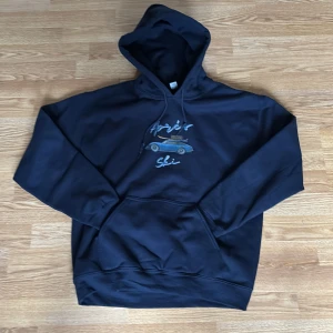 Svart hoodie med tryck 'Après Ski' och bilmotiv - Svart hoodie med tryck av en blå sportbil med skidutrustning på taket och texten 'Après Ski' på bröstet. Tröjan har huva och magficka, perfekt för en avslappnad stil.