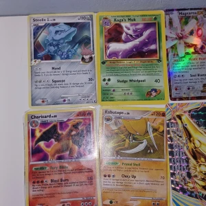 Pokémonkort samling med flera holografiska kort - Säljer en samling Pokémonkort med bland annat Steelix, Koga's Muk, Magearna, Charizard, Kabutops, Dialga, Hitmon-Top, Probopass och Skuntank. Flera av korten är holografiska och har olika färger och motiv. Perfekt för samlare eller Pokémonfans.