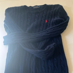 Mörkblå kabelstickad tröja från Polo Ralph Lauren - Mörkblå kabelstickad tröja från Polo Ralph Lauren med klassisk röd broderad logga på bröstet. Tröjan har rund halsringning och långa ärmar. Perfekt för lager-på-lager och stilren look. Scick 8/10 då knappt använd priset är ej hugget i sten