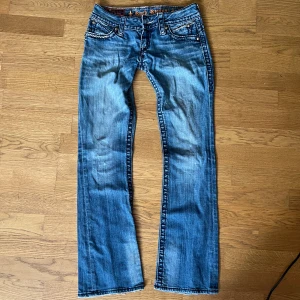 Blå jeansbyxor från Rock Revival - Säljer ett par blå jeansbyxor från Rock Revival med slitna detaljer och kontrastsömmar. Byxorna har klassisk femficksdesign, dekorativa knappar och broderad logga på insidan av linningen. Passformen är low waist med bootcut-stil. Måten ser man på bilderna. Priset kan diskuteras 