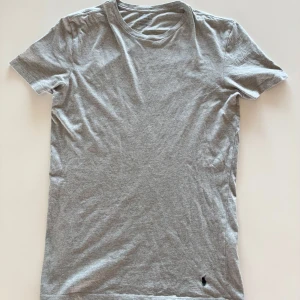 Slim fit Ralph Lauren T-Shirt - Säljer en klassisk grå t-shirt från Polo Ralph Lauren med liten broderad logga i mörkblått vid nederkanten. T-shirten är i mjuk bomull och har normal passform med korta ärmar. Perfekt basplagg till garderoben!
