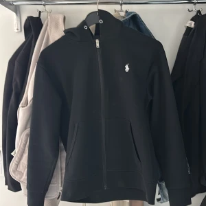 Svart hoodie med dragkedja från Polo Ralph Lauren - Svart hoodie från Polo Ralph Lauren med dragkedja framtill och vit broderad logga på bröstet. Tröjan har huva och två fickor framtill. Perfekt för en avslappnad stil. Säljer för att jag tröttnat och lite liten. Skick 10/10