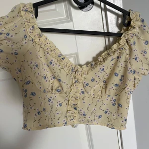 Gul blommig croppad blus med volang - Söt croppad blus i ljusgul färg med blått och rosa blommönster. Blusen har kort puffärm, volangdetaljer längs urringningen och knäppning framtill. Perfekt för vår och sommar!
