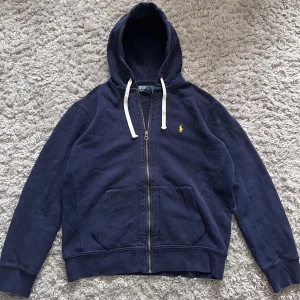 Marinblå hoodie från Polo Ralph Lauren - Säljer en marinblå hoodie från Polo Ralph Lauren med dragkedja och vit snörning i huvan. Tröjan har broderad gul logga på bröstet och två fickor framtill. Perfekt för en avslappnad stil. Mått: längd från axel: 61cm armhåla till armhåla:61 cm
