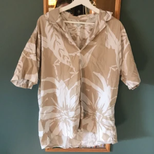 Beige och vit blommig skjorta - Säljer en beige skjorta med stora vita blommor och korta ärmar. Skjortan har klassisk krage och knappar framtill, perfekt för en avslappnad stil. Luftigt material som passar bra till varma dagar.