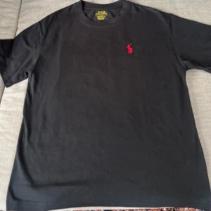 Svart t-shirt från Polo Ralph Lauren - Säljer en svart t-shirt från Polo Ralph Lauren med klassisk röd broderad logga på bröstet. T-shirten har rund halsringning och normal passform. Perfekt för en stilren och enkel look.