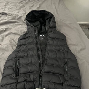 Svart dunväst med huva från Zara - Säljer en svart dunväst från Zara med huva och dragkedja framtill. Västen har två fickor med dragkedja och quiltad design. Perfekt för lager-på-lager under kyliga dagar.
