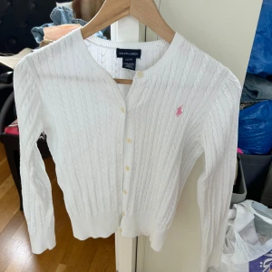 Vit kabelstickad kofta från Ralph Lauren - Vit kabelstickad kofta från Ralph Lauren med rund halsringning och knappar framtill. Klassisk design med ribbade muddar och Ralph Laurens rosa logga broderad på bröstet. Perfekt för lager-på-lager-stil. Storlek L (12/14) år barn tjej avdelningen men passar som xs på dam.