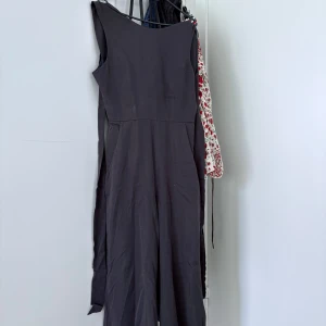 Grå lång jump suit - Säljer en grå jump suit med ärmlös design och rund halsringning. Jump suit har dekorativa knappar med metallöglor på axlarna och en stilren, minimalistisk look. Perfekt för dig som gillar enkel men snygg stil.