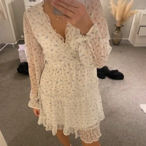 Vit blommig volangklänning med långa ärmar - Aldrig använd, Romantisk vit klänning med små blå blommor, volangdetaljer och v-ringning. Långa, transparenta ärmar och markerad midja. Perfekt för vår och sommar. Lätt och luftig känsla.