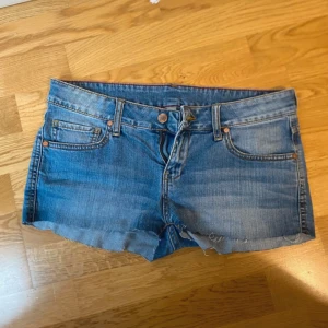 Low waisted Blå jeansshorts - Snygga blå jeansshorts med klassisk femficksdesign och rå, fransig kant nedtill. Perfekta för varma dagar och enkel att matcha med olika toppar. Stängs med knapp och dragkedja framtill.