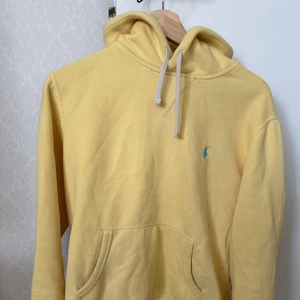 Gul hoodie från Polo Ralph Lauren - Gul hoodie från Polo Ralph Lauren med klassisk känguruficka och huva med vita snören. Liten blå logga broderad på bröstet. Perfekt för en avslappnad och stilren look.
