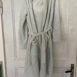 Ljusblå fleece morgonrock med huva från KappAhl - Mysig ljusblå morgonrock i fleece från KappAhl. Har huva med två fluffiga bollar och knytskärp i midjan. Långärmad och med fickor framtill. Perfekt för att hålla sig varm hemma.