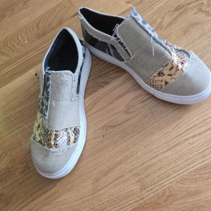 Beige slip-on sneakers med leopard- och ormmönster - Säljer ett par unika slip-on sneakers i beige canvas med detaljer i leopard- och ormmönstrad syntet. Skorna har vit platt sula och råa kanter för en cool look. Perfekta för dig som vill sticka ut med dina skor!