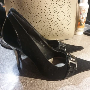 Svarta pumps med metallklack och dekordetalj - Säljer ett par eleganta svarta pumps med spetsig tå och hög metallklack. Skorna har en snygg metallspänne framtill och är tillverkade i skinn med blanka detaljer baktill. Perfekta för dig som vill sticka ut.