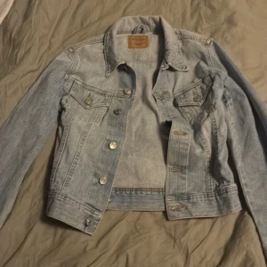 Ljusblå jeansjacka från Rocky - Säljer en klassisk ljusblå jeansjacka från Rocky med silvriga knappar och två bröstfickor med lock. Jackan har en tidlös design och är lätt att matcha till olika outfits. Perfekt för dig som gillar en avslappnad och trendig stil.