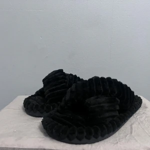 Svarta fluffiga sandaler - Mysiga svarta sandaler med korsade band och ribbad struktur. De har en platt sula och är tillverkade i ett mjukt, plyschliknande material som känns riktigt skönt mot foten. Perfekta att glida runt i hemma eller när du vill ha något bekvämt på fötterna.