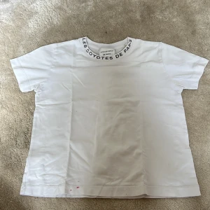 Vit t-shirt från Les Coyotes de Paris - Vit t-shirt med svart text 'LES COYOTES DE PARIS' runt halsringningen. Klassisk passform och rund hals. Tillverkad i mjuk bomull. Den har en nagellacks fläck.