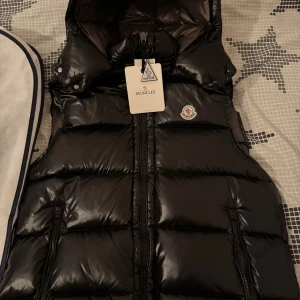 Svart dunväst från Moncler - Säljer en svart, glansig dunväst från Moncler med huva och dragkedja framtill. Västen har Moncler-logga på bröstet och två fickor med dragkedja. Perfekt för kyligare dagar och riktigt snygg att styla till olika outfits.