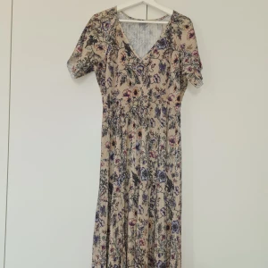 Blommig långklänning från Indiska - Säljer en romantisk långklänning från Indiska med blommigt mönster i lila, rosa och grönt på beige botten. Klänningen har v-ringning, korta ärmar och ett luftigt fall. Perfekt för sommarens alla tillfällen!
