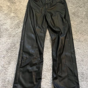 Svarta skinnbyxor från H&M - Säljer ett par svarta skinnbyxor från H&M med raka ben och klassisk passform. Byxorna har fickor bak och stängs med dragkedja och knapp framtill. Perfekta för en trendig look.