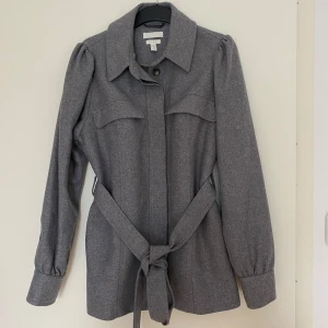 Grå kappa med bälte från H&M - Snygg grå kappa från H&M med markerad krage och knytbälte i midjan. Jackan har långa ärmar med knappar vid ärmslut och två bröstfickor. Perfekt för höst och vår när du vill ha en stilren look.