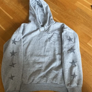 Grå hoodie med stjärnor från Gina Tricot - Grå hoodie från Gina Tricot med tryckta svarta stjärnor längs båda ärmarna och en stor stjärna samt texten 'MA CHÉRIE' på bröstet. Mjuk insida och klassisk huva. Perfekt för en avslappnad stil.