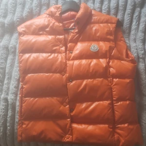 Orange dunväst från Moncler storlek 1 - Säljer en orange dunväst från Moncler med hög krage och tryckknappar framtill. Västen har Moncler-logga på bröstet och är vadderad med dun för extra värme. Perfekt för lager-på-lager under kyliga dagar