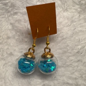 Glittriga örhängen med blå stjärnor - Unika örhängen med glaskulor fyllda av blått glitter och små stjärnor. Krokar och detaljer i guldfärgad metall. Perfekta för att ge din stil en färgglad och lekfull touch. Krokarna är i sterling silver