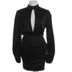 Svart kortklänning med lång ärm från Bianca x Nelly - Snygg svart kortklänning från Bianca x Nelly med lång ärm och hög hals. Klänningen har en djup v-ringad öppning framtill och markerad midja. Tillverkad i polyester med lite stretch för en smickrande passform.