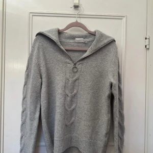 Grå stickad tröja med dragkedja  - Säljer en grå stickad tröja från vila med  kabelstickning framtill och dragkedja med ringdetalj. Tröjan har lång ärm och hög krage som kan vikas ner. Perfekt för kyliga dagar!