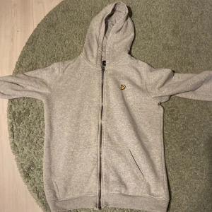 Grå hoodie från Lyle & Scott  - Säljer en grå hoodie från Lyle & Scott Junior med dragkedja framtill och huva. Tröjan har två fickor och en liten gul logga på bröstet. Perfekt för en avslappnad stil.
