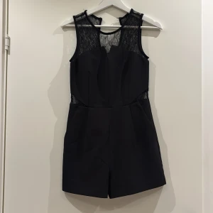 Svart ärmlös playsuit med spetsdetaljer - Säljer en svart ärmlös playsuit med snygga spetsdetaljer upptill och i midjan. Modellen har rund halsringning och markerad midja, vilket ger en stilren och modern look. Perfekt för dig som vill ha något elegant men ändå bekvämt.