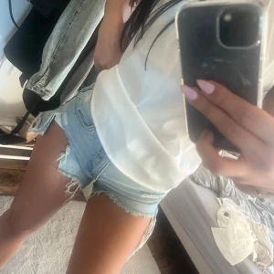 jeansshorts - Säljer ett par ljusblå jeansshorts med råa franskantsdetaljer och slitningar. Klassisk femficksmodell med låg midja och kort passform. Perfekta för varma dagar och enkel att matcha med olika toppar.