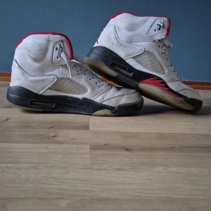  Air Jordans 5 vita med röda detaljer - Säljer ett par klassiska  Air Jordans 5 i vitt med röda detaljer och svart sula. Skorna har snörning, meshpaneler på sidorna och rund tå. Perfekta för dig som gillar retrostil och streetwear.