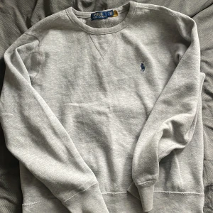 Grå sweatshirt från Polo Ralph Lauren - Klassisk grå sweatshirt från Polo Ralph Lauren med rund halsringning och broderad blå logga på bröstet. Tröjan har långa ärmar och ribbade muddar vid ärmslut och nederkant. Perfekt för en avslappnad stil.