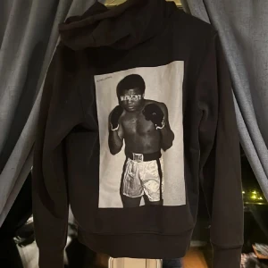 Svart hoodie med boxningsmotiv - Svart hoodie med stor svartvit bild av en Muhammed Ali på ryggen. Tröjan har huva med dragsko och en stor magficka framtill. Ny pris 600kr