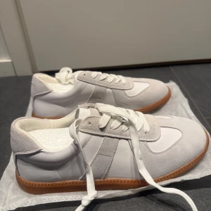 Maison Margiela - Säljer ett par stilrena sneakers i vitt och ljusgrått med snygga mockadetaljer och klassisk brun sula. Skorna har snörning och en rund tå, perfekta för en clean och modern look. Storleken passar både 40 _41. Använda några gånger 