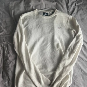 Vit stickad tröja från GANT - Säljer en klassisk vit stickad tröja från GANT i premium bomull. Tröjan har rund halsringning, diskret logga på bröstet och långa ärmar. Perfekt för en stilren och enkel look.