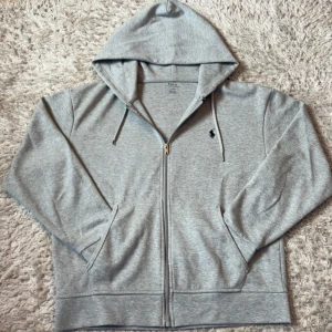 Grå hoodie från Polo Ralph Lauren - Säljer en klassisk grå hoodie från Polo Ralph Lauren med dragkedja och huva. Tröjan har långa ärmar, två fickor framtill och en liten broderad logga på bröstet. Perfekt för en avslappnad stil.