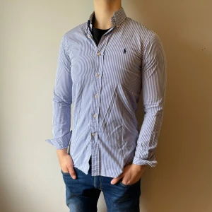 Randig Ralph lauren skjorta - Stilig skjorta från Ralph lauren med blåa och vita ränder och en marinblå logga, skicket är mycket bra överlag förutom en liten fläck längst ner vid magen men det är inget som syns storleken är 14 1/2 den är 70 cm lång och passar storlek s, tveka inte att höra av dig om du har frågor!