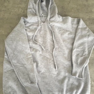 Grå hoodie med huva och snörning - En basic grå hoodie med huva och snörning framtill. Tröjan har långa ärmar och ribbade muddar vid ärmslut och nederkant. Perfekt för chill dagar eller när du vill ha en enkel och stilren look.