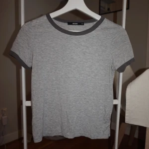 Grå t-shirt med kontrastkant Bik Bok - Enkel och stilren ljusgrå t-shirt från Bik Bok med mörkgrå kontrastkant vid halsringning och ärmslut. Klassisk passform och korta ärmar, perfekt till jeans eller kjol. Mjuk och skön bomullskänsla.