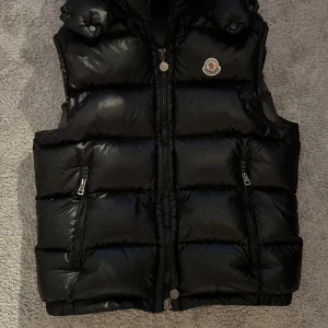 Svart dunväst från Moncler - Snygg svart dunväst från Moncler med glansig finish och klassisk logga på bröstet. Västen har två dragkedjefickor framtill och hög krage. Perfekt att slänga över en hoodie eller tröja för en cool look.