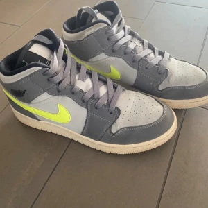 Nike Air Jordan 1 Mid grå/gul Gunsmoke Volt - Nike Air Jordan 1 Mid sneakers i Gunsmoke Volt. Klassisk high-top siluett med snörning, perforerad tå och vit sula. Skorna har svart logga på sidan och svart/vit etikett på plösen. 