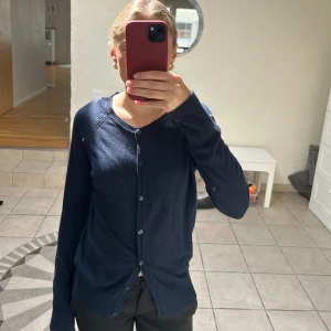 Mörkblå kofta från Name It - En stilren mörkblå kofta från Name It med rund halsringning och knappar hela vägen framtill. Lätt och tunn modell med långa ärmar och ribbade muddar. Perfekt för lager-på-lager och enkel att matcha med det mesta.